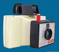 Side angle view of the Polaroid Swinger Model 20 camera showing cream body, black viewfinder, and front lens with red lever.