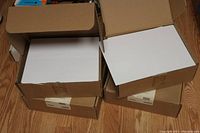 Four unopened cardboard boxes filled with blank white labels stack on wooden floor.