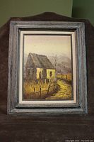 Frontal photo of the painting showing the farmhouse barn autumn scene contained in a weathered blue-gray wooden frame on a brown fabric backdrop.