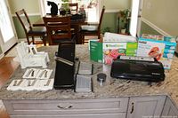Wide shot showing all items in the lot arranged on kitchen countertop including the white KitchenAid spiralizer, black KitchenAid mandoline slicer, FoodSaver vacuum sealer, and two boxes of sealing bags.