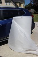 Full view of large roll of white polyethylene foam standing next to a blue vehicle for scale, with some foam unfurled on the ground.