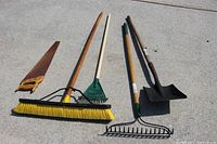 Photo of garden tools laid on a concrete surface showing a hand saw, a push broom, a green leaf rake, a garden hoe, a shovel, and a garden rake.
