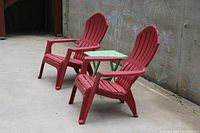 Two red Adirondack chairs with a small green foldable table between them, placed on concrete against a chalk-drawn wall.