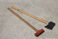 View of sledge hammer with red head and axe with black head, both with wooden handles on concrete ground.