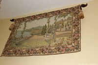 Full view of the large tapestry hanging on a wall, showing the floral border and scenic garden image with patio, bench, trees, water, and architectural features.