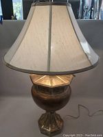 Front view of the lamp showing the gold gilded base with geometric design and beige fabric lampshade.