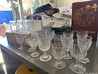 Overview of lot showing assortment of small antique glasses on a kitchen counter. Includes small wine/cordial glasses with etched and cut glass designs, and clear vintage jelly jars in the background.