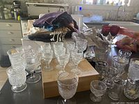 Photo shows a variety of clear glass mini wine glasses, cordials, goblets, and a few red tinted glasses on a kitchen counter with other household items in background.