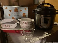 Photo showing the red baking dish with lid, three Corning Ware Rosemarie casserole dishes, heart-shaped non-stick mini cake pans box, vintage 2-cup measuring cup, and Instant Pot cooker on a shelf.