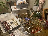 Wide view of trays and table with assorted costume jewelry including necklaces, bracelets, earrings, and watches laid out, showing large quantity and diversity of pieces.