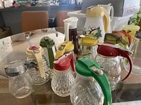 Overview of all two vintage pitchers and twelve syrup bottles with various colored plastic tops arranged on glass table.