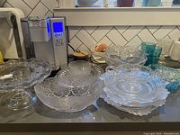 Top view of various clear glass antique bowls, plates and serving pieces arranged on a kitchen counter showing detailed pressed glass floral and geometric patterns, including footed cake plate and handled bowl.