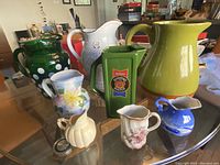 Lot of nine ceramic pitchers displayed on glass table, showing a variety of styles, colors, and shapes including polka dot, solid color, floral, and advertising designs.