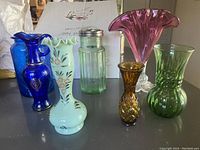Full group photo showing all six vases and the light green glass Avon decanter. Shows variety of colors: cobalt blue, pale green, amber, pink, green. Confirms no visible chips or cracks.