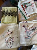 Photo shows a collection of embroidered vintage tea towels with motifs including a dancing figure at piano and a cartoonish animal playing an instrument, some towels have spots and stains visible.