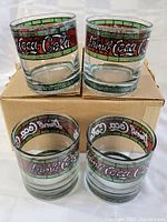 Four clear Coca-Cola drinking glasses featuring colored stained glass style bands with 'Drink Coca-Cola' text, displayed on and near a brown cardboard box.