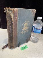 Full view of antique hardcover book showing blue cloth cover with gold embossed portrait and signature, alongside a water bottle for size reference. Spine damage visible.