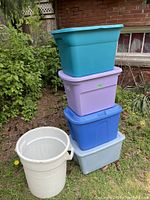Photo showing four rectangular plastic storage totes stacked outside, colors teal, purple, blue, gray, and one white cylindrical plastic garbage can next to them.