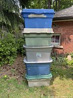 Five large plastic storage totes stacked outdoors against a brick wall and greenery. Visible colors include blue, beige, green, and gray, all with lids. Some wear and scuffs are visible on the totes consistent with used condition.