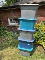 Stack of 5 plastic storage totes of different colors with lids seen outdoors against a brick wall and shrubbery.
