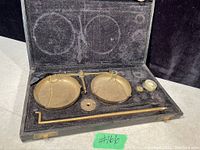 Photo showing antique solid brass balance scale with two pans inside original black fabric-lined case along with partial set of brass weights and balance arm rod, showing age wear and some missing weights.