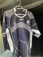 Front view of the men's Rugby Nations Scotland rugby shirt hanging on a hanger showing the navy and white striped design with logos embroidered on chest.