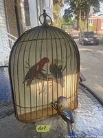 Full view of vintage metal bird cage wall art showing painted bird scene inside and ceramic bird figure attached to cage base outside.