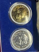 Gold and silver Ronald Reagan 35th Anniversary commemorative coins showing profile side in protective cases on blue velvet interior of display box.