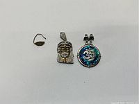 Three jewelry items: a small broken gold ring, silver figurative pendant with stones, and silver pendant with abalone shell and Om symbol