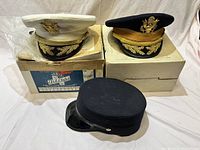 Three service hats displayed on and near two cardboard boxes. White and navy peaked caps with embroidered gold details and eagle insignia plus one black flat-topped cap with glossy visor.