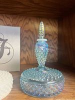 Iridescent blue Fenton hobnail textured glass bottle with pointed stopper displayed on wooden shelf, showing the bottle's body, lid, and stopper in excellent condition.