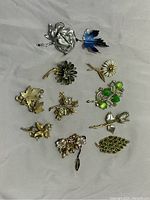 Lot of 10 floral-themed vintage pins on gray fabric background showing various metal tones and enamel work.