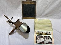 Photo showing the vintage wooden and metal handheld stereoscopic picture viewer along with a large stack of stereoscopic camera cards with printed images.
