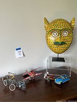 Yellow wooden monkey mask with green dotted pattern on display alongside five miniature models made from recycled drink cans: three bicycles (green, yellow, blue), one tricycle, one airplane, and one car.