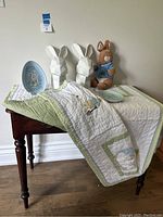 Lot displayed on wooden table with Pottery Barn Kids comforter blanket, two white geometric bunny faces, Peter Rabbit soft toy, and two decorative plates. Lighting shows details of embroidered blanket and color of soft toy.