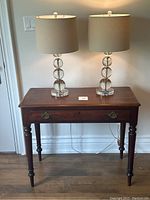 Two modern crystal ball table lamps lit on a wooden table with beige lamp shades, showing front view and cords visible.