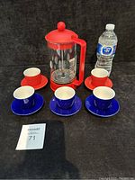 Six Nespresso espresso cups (three red, three blue) each with matching saucers, arranged around a Bodem French press plunger with red accents, all placed on black fabric background with water bottle for scale.