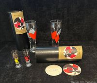 Two Ritzenhoff beer glasses each decorated with vertical red hearts and abstract black and white shapes, placed beside their original black cylindrical boxes with Ritzenhoff labeling. Two small shot glasses with cartoon character designs and two round coasters lay in front.