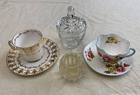 Two bone china teacups with matching saucers, a cut glass sugar dish with lid, and a heavy glass candle holder arranged on a light cloth surface