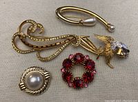 Four gold-tone costume brooches arranged on beige background: bird with pearl tip, curved pin with pearl, red rhinestone circle, faux pearl circle