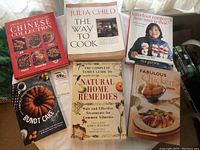Seven cookbooks arranged in two rows on a wooden floor with natural light and curtain background.