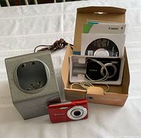 Photo showing entire lot with Casio Exilim digital camera in red, Canon PowerShot A800 camera in box with accessories, and vintage Brumberger slide viewer.