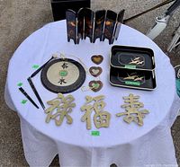 Photo of large round dinner gong on white tablecloth along with lacquered trays, small folding screen, heart-shaped picture frames, brass trivets with Chinese characters, and chopsticks arranged around it.