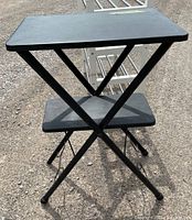 Front view of the black collapsible 2-tier metal stand with crossbar legs, placed outside showing ground
