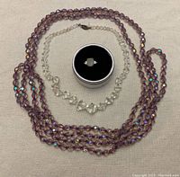 Two sparkly beaded necklaces with one having a clasp and a sterling silver ring with moonstone displayed in a circular black ring box.