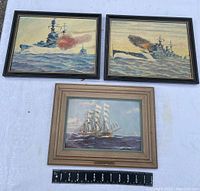 Group photo of the three framed nautical paintings: two battleships firing and one sailing ship titled 'The Garthpool'. The artworks are displayed with a measuring scale for size reference.