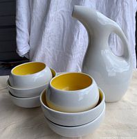 Photo showing white ceramic pitcher and stack of 7 ceramic bowls with yellow and white interiors.