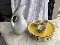 White ceramic pitcher with unique modern round handle next to stacked yellow dinner plates and three small white ramekins with yellow exterior ribbing on a beige tablecloth.