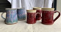 Five pottery mugs arranged on cloth backdrop, three red signed mugs and two blue-purple drip glaze mugs.