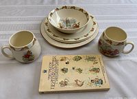 Photo shows two Bunnykins cups, two plates, one bowl stacked, and a Beatrix Potter book in front, displaying lot contents.